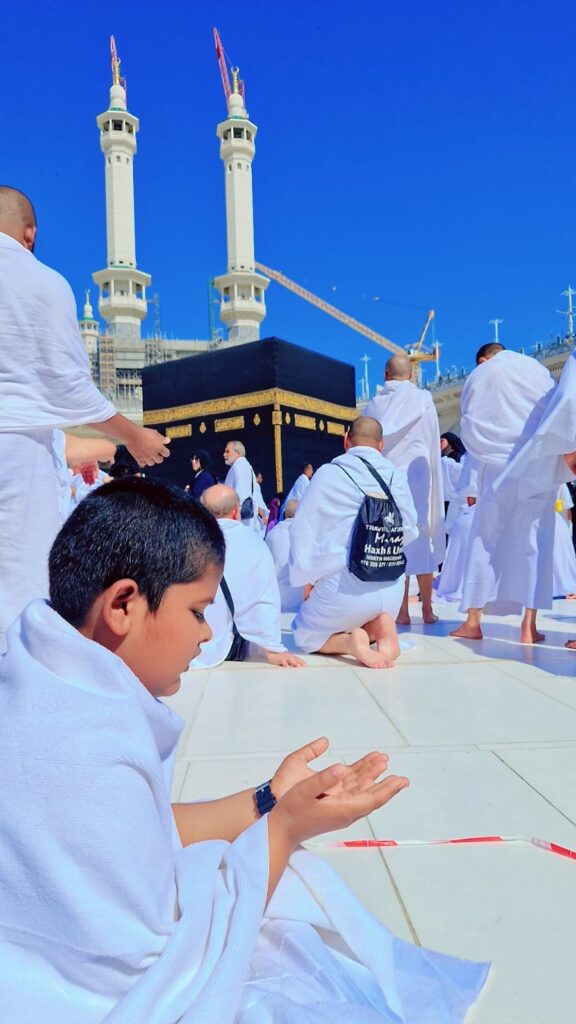 Young boys praying near the Kaaba in Mecca, with worshippers gathered around in a spiritual setting.